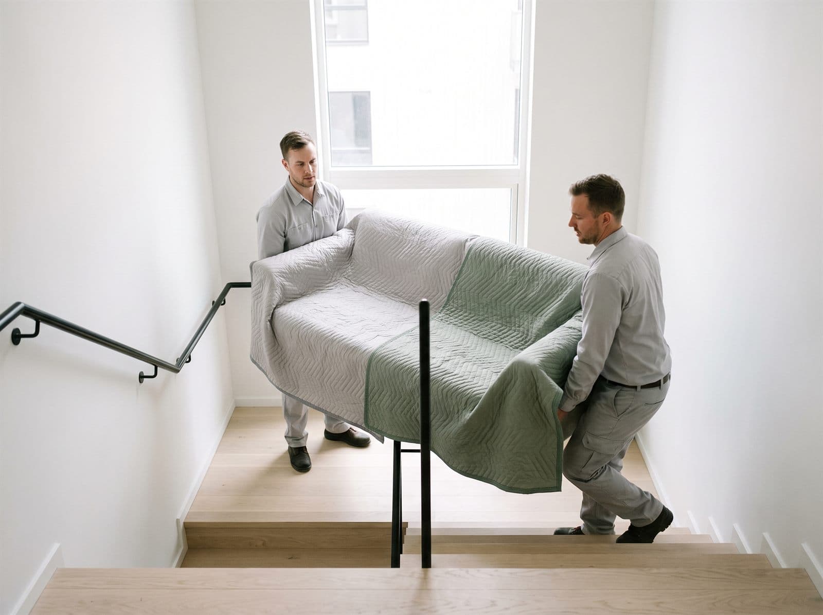 Två flyttare bär en inklädd soffa i ett ljust trapphus